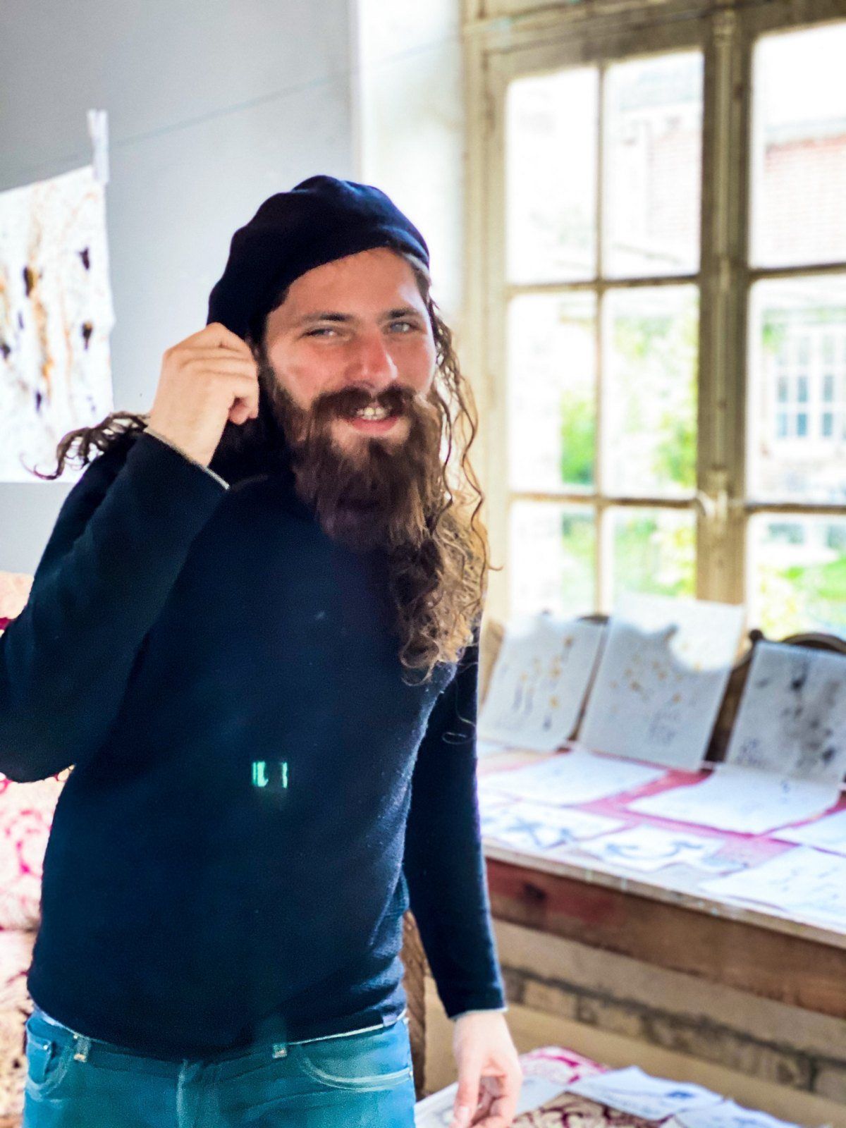 Aaron in a beret, smiling, in a sunlit studio with ink works on a bench