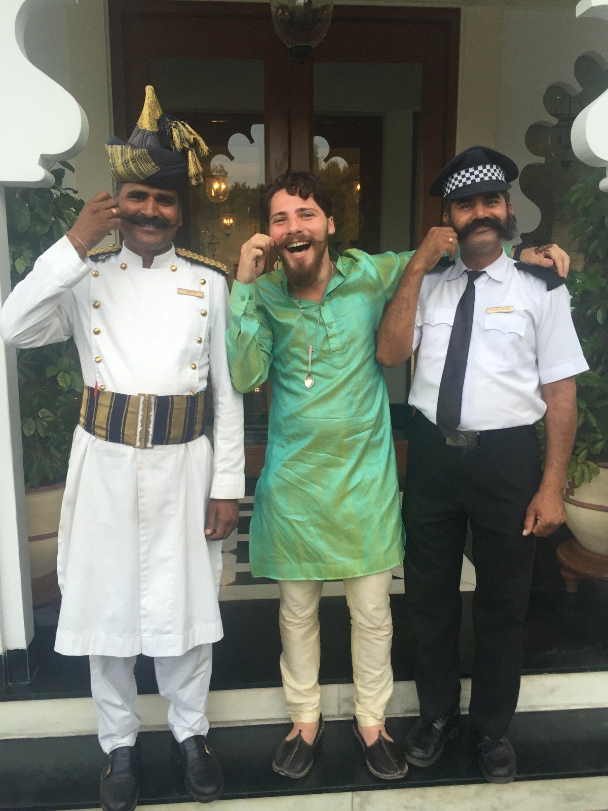 Aaron in a green kurta, twirling his moustache with two doormen at a hotel entrance