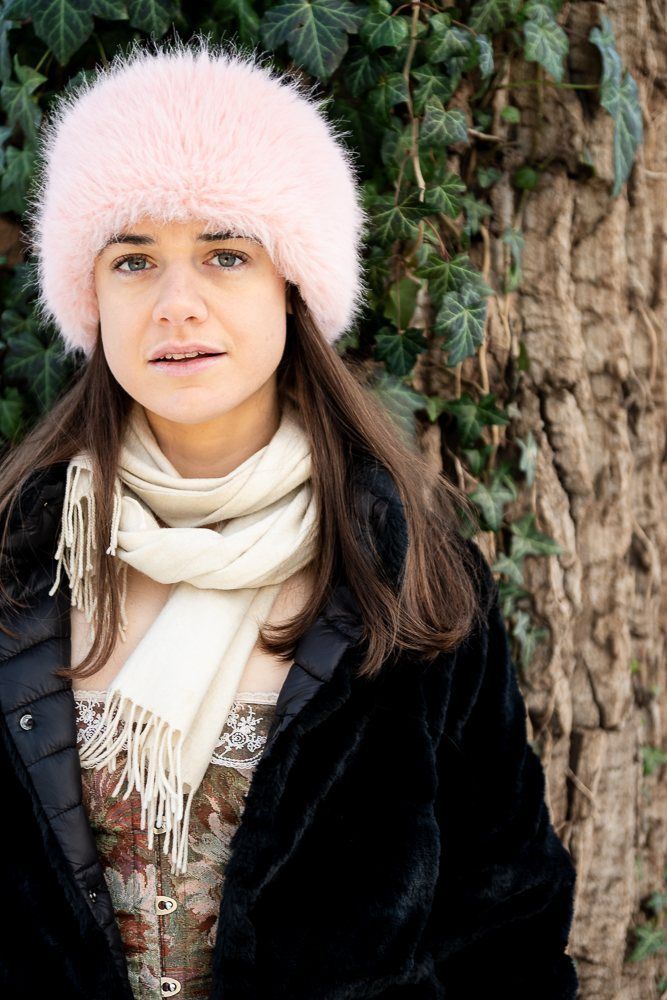 Julia in a pink fur hat against a tree trunk