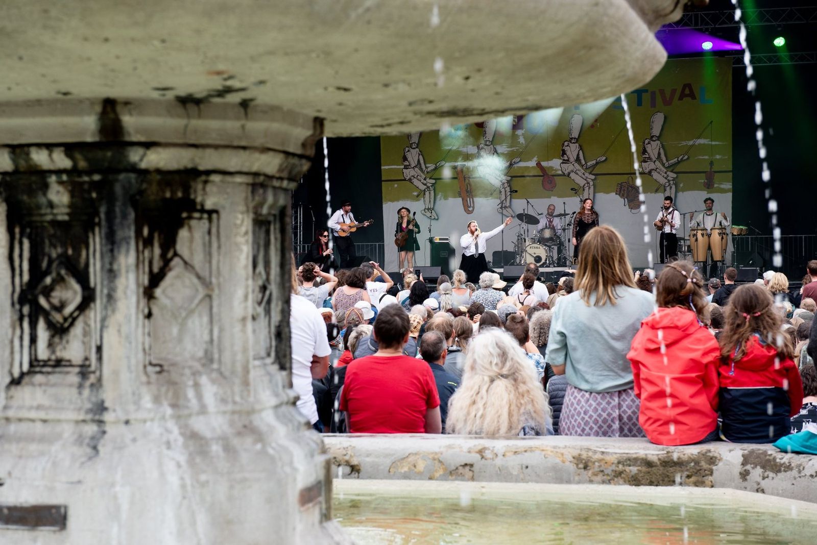 The band on stage seen past the stone fountain in the market square, the audience in front