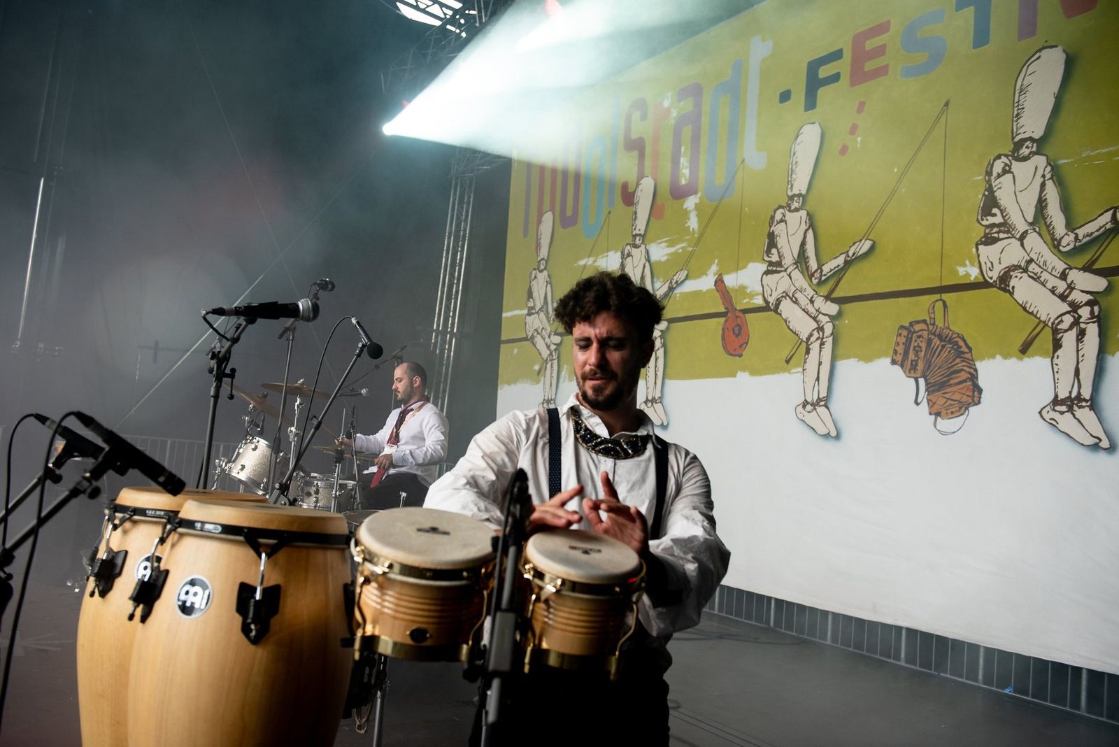 The percussionist on congas under a spotlight, the festival banner behind him